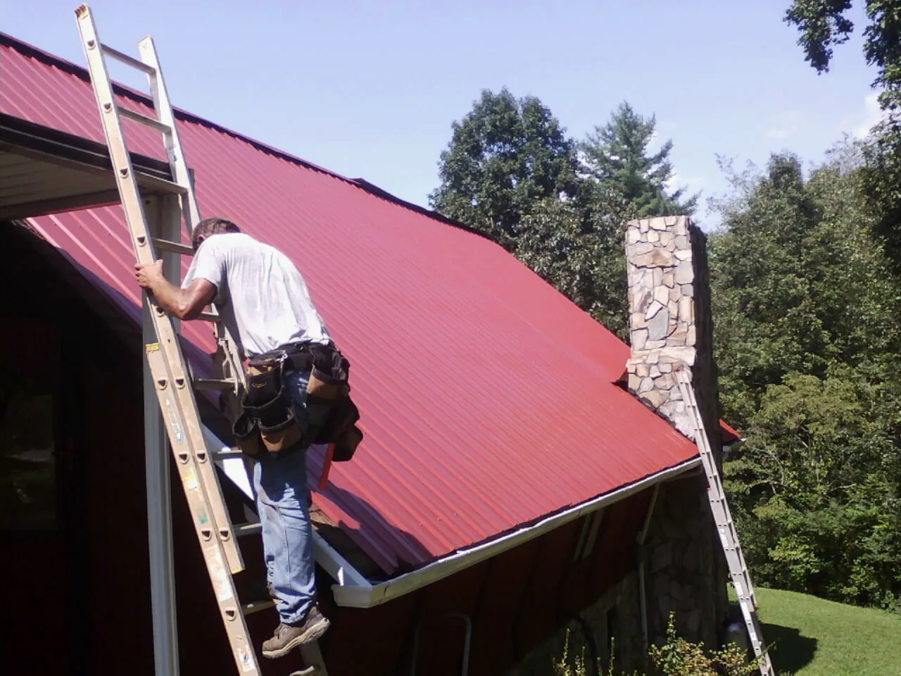 Roofer on a ladder beside a red metal roof and stone chimney — Soffit & Fascia Repair by Roofing Brothers in Quarryville